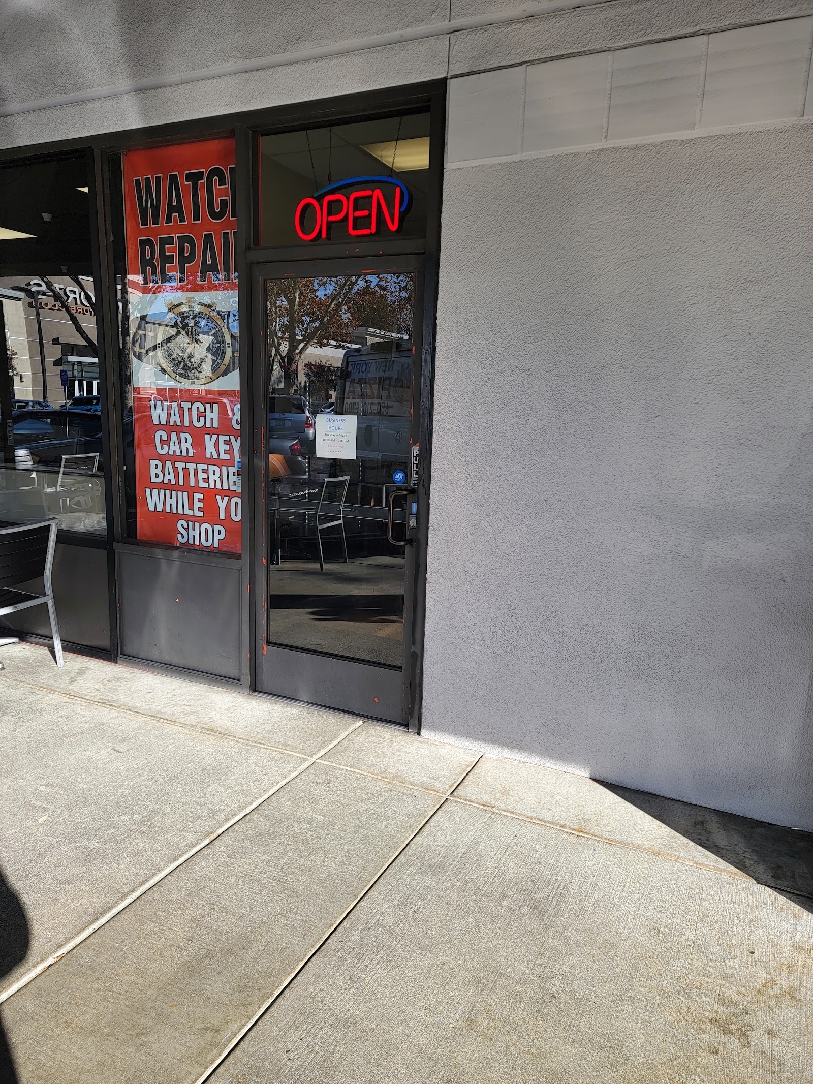 The shop door at M and S Watch Gallery in Grant Road Plaza, with a red neon OPEN sign and a poster reading watch and car key batteries while you shop.
